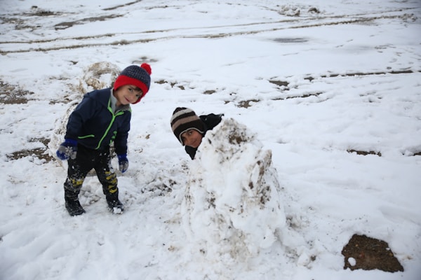 A child dressed in winter clothing stands on a snowy ground next to a snow mound. The child is wearing a blue jacket, red and blue knit hat, and gloves, with snow covering parts of their clothing and boots. Another person, partially visible, appears to be hiding behind the snow mound. The setting is a snow-covered area with some brown patches of ground visible.