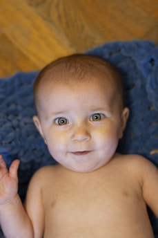 A baby is lying on a soft, textured blue blanket. The baby has a cheerful expression, with green eyes, and is looking directly at the camera. The background features a wooden floor.
