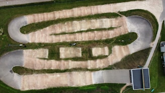 An aerial view of a dirt track designed for BMX or motocross racing. The track includes multiple lanes with bends and straight sections separated by grassy areas. It features banked turns and mounds typically used for jumps or obstacles. Surrounding the track is a green lawn, and there is a paved area with a shelter or small building to the side.