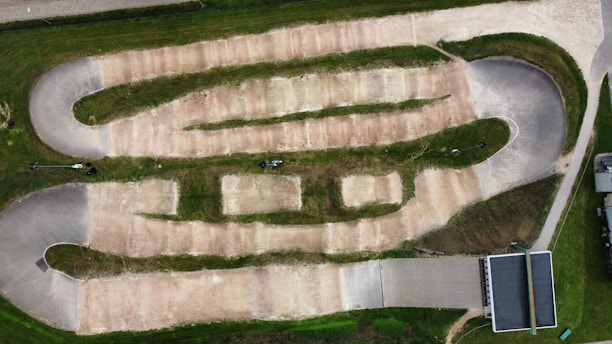 An aerial view of a dirt track designed for BMX or motocross racing. The track includes multiple lanes with bends and straight sections separated by grassy areas. It features banked turns and mounds typically used for jumps or obstacles. Surrounding the track is a green lawn, and there is a paved area with a shelter or small building to the side.