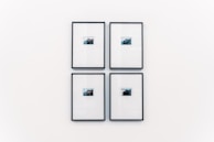 Four small framed photographs are arranged in a grid on a plain white wall. Each photograph features a landscape with water and a distant structure, possibly an island or mountain. The images are set in minimalistic black frames with a clean and modern aesthetic.