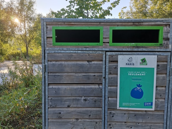 A wooden recycling or waste disposal container is situated outdoors with two rectangular openings framed in green. A sign with the Paris logo and instructions is attached to it. The background features lush green trees and shrubbery, with sunlight filtering through the leaves.