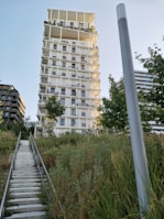 A modern, multi-story apartment building with a unique architectural design featuring an irregular pattern of contrasting light and dark sections. The building is surrounded by lush greenery and the sky is clear. A staircase ascends through grassy foliage leading up to the structure.