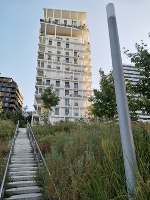 A modern, multi-story apartment building with a unique architectural design featuring an irregular pattern of contrasting light and dark sections. The building is surrounded by lush greenery and the sky is clear. A staircase ascends through grassy foliage leading up to the structure.