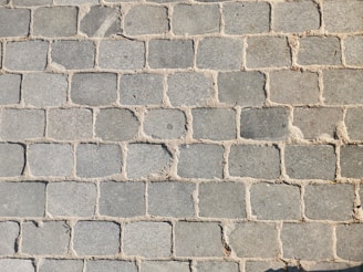 A pattern of rectangular stone pavers arranged in a grid, with varying shades of gray and beige grout filling the gaps between the stones.