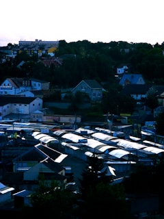 A townscape with clustered houses and a series of market stalls lined up, covered with white tarps. Vegetation and trees surround the area, while a hill in the distance supports various buildings.