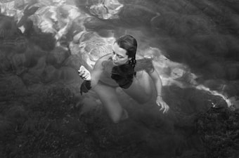 A person is submerged in clear water, surrounded by ripples and reflections on the water surface. Their expression appears contemplative as they examine a small stone or shell in their hand, with aquatic plants visible beneath.