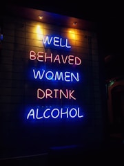 A neon sign with vibrant blue and pink letters displays the phrase 'Well behaved women drink alcohol' against a dark background. The text is arranged vertically across four lines, creating a striking visual contrast.