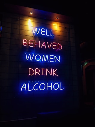 A neon sign with vibrant blue and pink letters displays the phrase 'Well behaved women drink alcohol' against a dark background. The text is arranged vertically across four lines, creating a striking visual contrast.