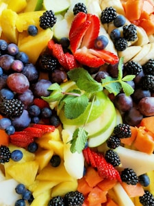 A vibrant assortment of fresh fruits arranged closely together, including strawberries, blackberries, blueberries, grapes, green apple slices, pineapple chunks, banana slices, papaya, and a sprig of mint at the center.