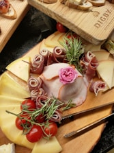 An assortment of cheeses, cured meats, and tomatoes arranged on a wooden board, garnished with fresh rosemary and a decorative pink flower. Slices of bread are positioned above the main board.