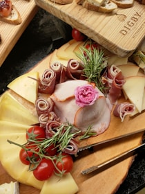 An assortment of cheeses, cured meats, and tomatoes arranged on a wooden board, garnished with fresh rosemary and a decorative pink flower. Slices of bread are positioned above the main board.