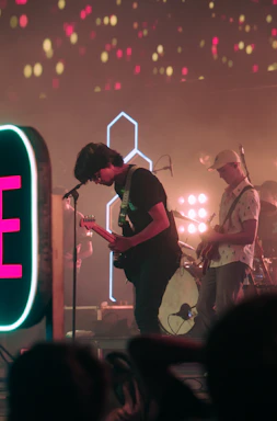 A band is performing on stage with vibrant lighting effects. Two musicians are in view, one playing a guitar and singing into a microphone while the other is also playing a guitar. The background features colorful lights and a large neon sign is visible on the side. The atmosphere suggests a lively, energetic concert setting.