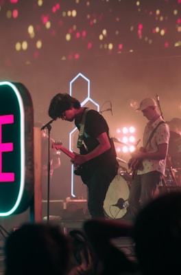 A band is performing on stage with vibrant lighting effects. Two musicians are in view, one playing a guitar and singing into a microphone while the other is also playing a guitar. The background features colorful lights and a large neon sign is visible on the side. The atmosphere suggests a lively, energetic concert setting.