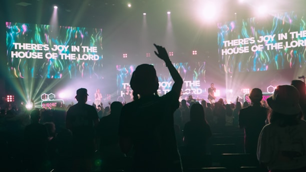 A lively concert scene with a crowd of people enjoying live music. The stage is brightly lit with colorful neon lights and multiple large screens displaying the text 'THERE'S JOY IN THE HOUSE OF THE LORD'. The audience appears engaged, with one person prominently raising their hand.