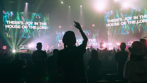A lively concert scene with a crowd of people enjoying live music. The stage is brightly lit with colorful neon lights and multiple large screens displaying the text 'THERE'S JOY IN THE HOUSE OF THE LORD'. The audience appears engaged, with one person prominently raising their hand.
