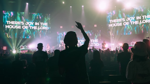 A lively concert scene with a crowd of people enjoying live music. The stage is brightly lit with colorful neon lights and multiple large screens displaying the text 'THERE'S JOY IN THE HOUSE OF THE LORD'. The audience appears engaged, with one person prominently raising their hand.