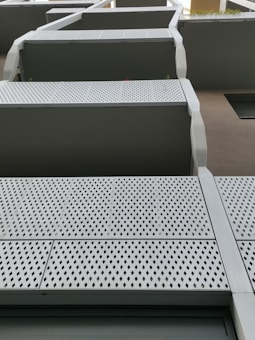 Modern architectural design featuring multiple levels of balconies. The balconies have perforated metal panels with a diamond pattern and are framed by sleek, metallic edges. The structure is part of a larger building that has a minimalist aesthetic.