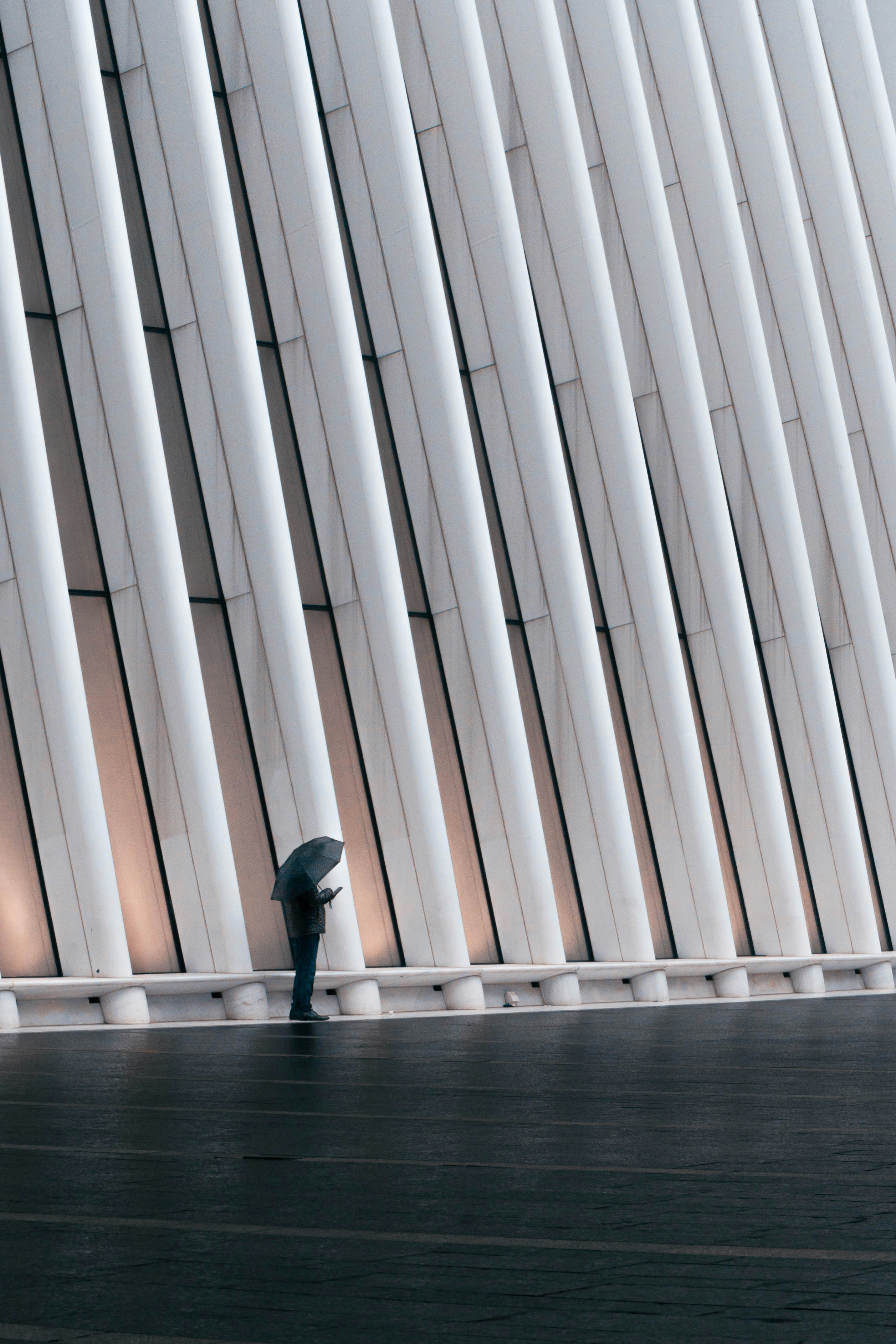 A photograph of a person standing alone in the rain against a modern building facade.