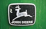 A metallic emblem with a leaping deer is centered on a green background. The deer is positioned inside a rectangular black frame with rounded corners. Below the deer image, the words 'John Deere' are printed in bold, capitalized white letters.