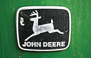 A metallic emblem with a leaping deer is centered on a green background. The deer is positioned inside a rectangular black frame with rounded corners. Below the deer image, the words 'John Deere' are printed in bold, capitalized white letters.