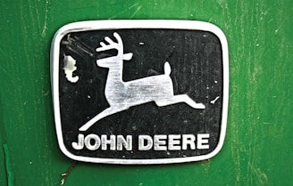 A metallic emblem with a leaping deer is centered on a green background. The deer is positioned inside a rectangular black frame with rounded corners. Below the deer image, the words 'John Deere' are printed in bold, capitalized white letters.