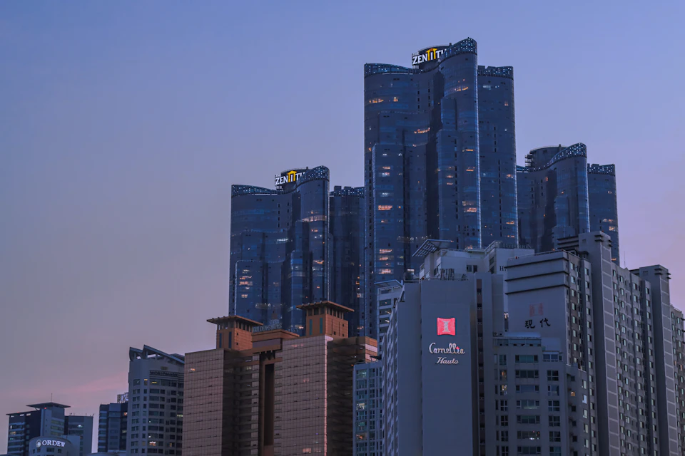 The image depicts a city skyline at dusk, with tall buildings illuminated against the darkening sky. The tallest building is a modern skyscraper with a curved facade and numerous windows that reflect the fading light. Below it are several other buildings, including one with a distinctive red sign on its facade. The colors in the scene are predominantly blue and gray, creating a serene atmosphere as the sun sets over the cityscape.