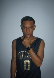 A person is standing against a plain background, wearing a black sleeveless jersey with gold lettering and the number zero. They have short hair and are accessorized with a watch, necklace, earrings, and a bracelet. The expression is contemplative, with a hand placed under the chin.