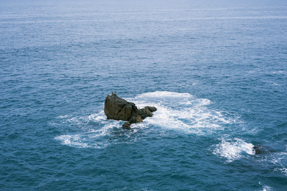 Large rock formation in deep blue ocean water with crashing white waves nearby