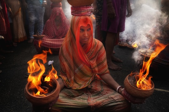 A person dressed in vibrant, colorful traditional attire is sitting on the ground during a religious ritual. They balance a pot on their head and hold two pots with open flames, surrounded by smoke. The person is part of a larger gathering, with others similarly dressed in the background participating in the event.