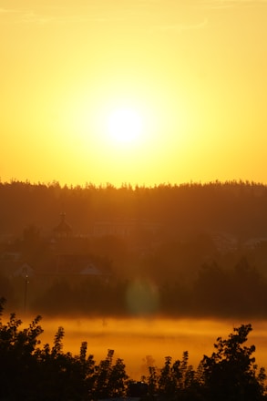 A vibrant sunrise illuminates a rural landscape, with a bright sun hovering above the horizon. The golden light creates a warm glow over the silhouetted trees and buildings in the distance.