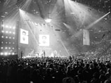 A large concert venue filled with a crowd of people. The stage is illuminated with bright spotlights, and a band or musical group is performing. Confetti fills the air, creating a lively and festive atmosphere. Two large screens display images, suggesting a celebration or tribute.