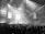 A large concert venue filled with a crowd of people. The stage is illuminated with bright spotlights, and a band or musical group is performing. Confetti fills the air, creating a lively and festive atmosphere. Two large screens display images, suggesting a celebration or tribute.