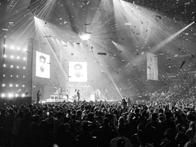 A large concert venue filled with a crowd of people. The stage is illuminated with bright spotlights, and a band or musical group is performing. Confetti fills the air, creating a lively and festive atmosphere. Two large screens display images, suggesting a celebration or tribute.