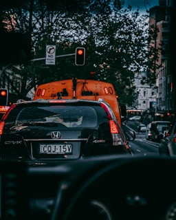 A busy urban street scene featuring cars lined up in traffic. A black Honda CR-V is in the foreground with a visible license plate. Red traffic lights are illuminated, and there are buildings and trees lining the street. A no right turn sign is also visible.