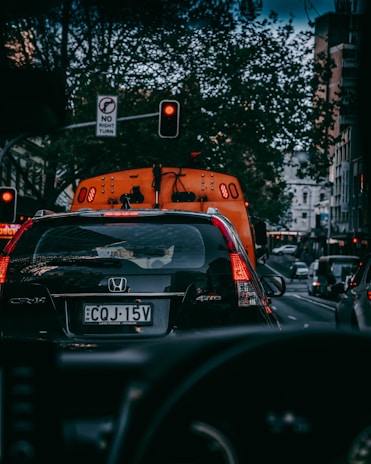 A busy urban street scene featuring cars lined up in traffic. A black Honda CR-V is in the foreground with a visible license plate. Red traffic lights are illuminated, and there are buildings and trees lining the street. A no right turn sign is also visible.
