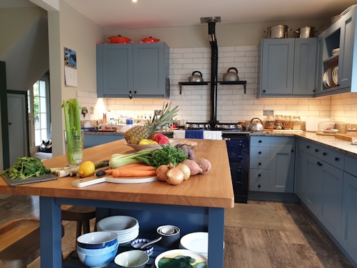A cozy kitchen featuring blue cabinets and a central wooden island. The countertop displays an array of fresh produce including carrots, celery, onions, potatoes, a pineapple, and a lemon. There's a vintage stove with kettles on the back wall, and the shelves are stocked with jars and dishes. The ambiance is warm and inviting.