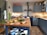 A cozy kitchen featuring blue cabinets and a central wooden island. The countertop displays an array of fresh produce including carrots, celery, onions, potatoes, a pineapple, and a lemon. There's a vintage stove with kettles on the back wall, and the shelves are stocked with jars and dishes. The ambiance is warm and inviting.