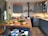 A cozy kitchen featuring blue cabinets and a central wooden island. The countertop displays an array of fresh produce including carrots, celery, onions, potatoes, a pineapple, and a lemon. There's a vintage stove with kettles on the back wall, and the shelves are stocked with jars and dishes. The ambiance is warm and inviting.