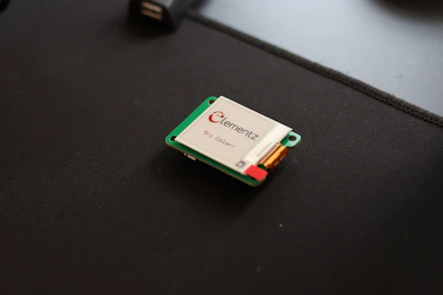 A small electronic device, likely a microcontroller or a sensor, is placed on a dark surface. It has a green circuit board and is labeled with 'Elementz.' There are visible components, including a small rectangular screen or panel.