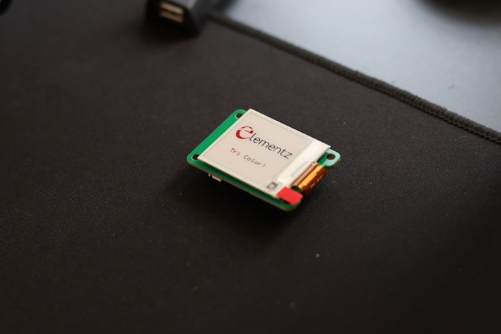 A small electronic device, likely a microcontroller or a sensor, is placed on a dark surface. It has a green circuit board and is labeled with 'Elementz.' There are visible components, including a small rectangular screen or panel.