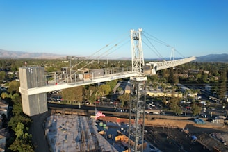 A large tower crane is prominently situated above a construction site, surrounded by a residential or suburban area. It is towering over the under-construction structure with a visible American flag attached to it. The backdrop features a sprawling landscape with a mix of trees, buildings, and distant hills under a clear blue sky.