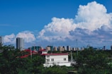A skyline featuring numerous high-rise buildings under a vibrant blue sky with large, fluffy clouds. In the foreground, trees and a few low structures with red and white rooftops are visible.