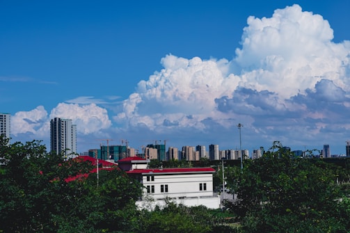 A skyline featuring numerous high-rise buildings under a vibrant blue sky with large, fluffy clouds. In the foreground, trees and a few low structures with red and white rooftops are visible.