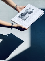 A pair of hands are holding a stack of printed papers with a cover depicting a black and white photograph of two people conversing. The paper is titled 'Annie Hall,' with several names listed alongside the image. The hands are adorned with colorful bracelets, and the scene is lit sharply, casting a prominent shadow on the ground.