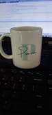 A white ceramic mug with the letter 'P' and the name 'Pravin' printed on it is placed on a keyboard. A computer screen displaying code is visible in the background.