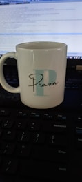 A white ceramic mug with the letter 'P' and the name 'Pravin' printed on it is placed on a keyboard. A computer screen displaying code is visible in the background.