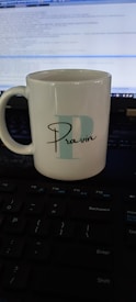 A white ceramic mug with the letter 'P' and the name 'Pravin' printed on it is placed on a keyboard. A computer screen displaying code is visible in the background.