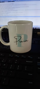 A white ceramic mug with the letter 'P' and the name 'Pravin' printed on it is placed on a keyboard. A computer screen displaying code is visible in the background.