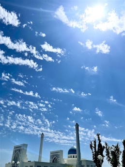 A mosque with tall minarets and a blue dome stands prominently against a backdrop of a bright blue sky filled with scattered white clouds. The sun shines brightly, casting a warm glow over the scene. There are trees to the side, adding greenery to the foreground.